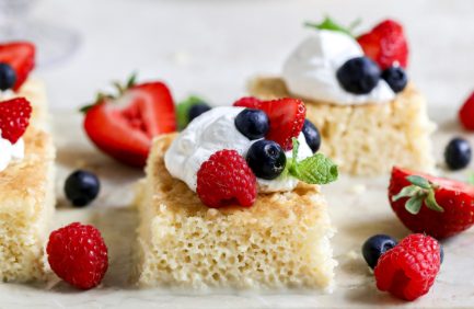 Sponge cake squares topped with whipped cream, fresh strawberries, blueberries, raspberries, and a mint leaf, served on a light-colored surface.