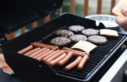 Hot dogs and hamburger patties, some topped with cheese, are cooking on a grill outdoors.