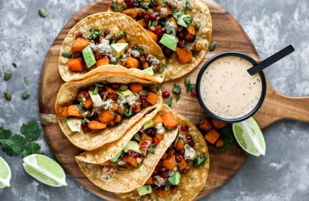 Four loaded vegetable tacos topped with avocado and herbs, served on a wooden board with a creamy dipping sauce and lime wedges on the side.