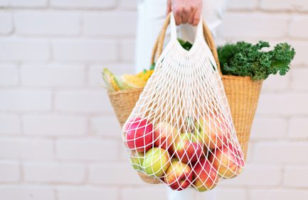 Girl holding mesh shopping bag full of apples and straw bag with organic vegetables, brick background. Zero waste, plastic free concept. Sustainable lifestyle. Copy space