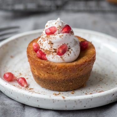 A small dessert tart topped with whipped cream, pomegranate seeds, and chocolate shavings, served on a white plate.