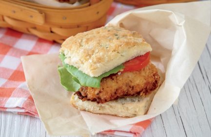A crispy fried chicken sandwich with lettuce and tomato on a biscuit, wrapped in parchment paper on a table.