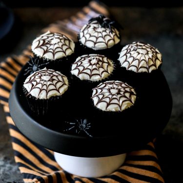 Six cupcakes decorated with white icing and black spiderweb designs on a black cake stand, with small plastic spiders and a striped cloth underneath.