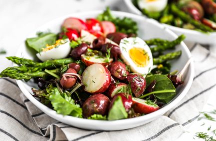 A fresh salad with leafy greens, radishes, olives, tomatoes, asparagus, and a halved soft-boiled egg served in a white bowl.