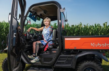 Laurie Hayn, an Indiana farmer that works with AgrAbility, sitting in the cab of a Kabota with a corn field in the background