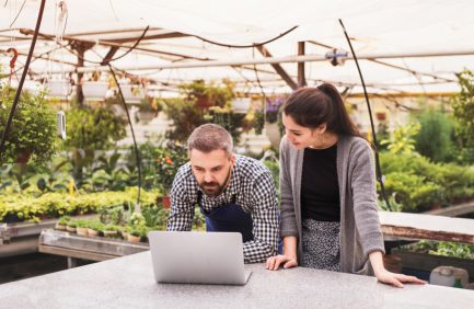 Young gardeners with laptop in a large greenhouse with pots of seedlings and plants.