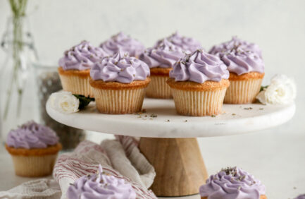 Vanilla cupcakes with purple frosting and sprinkles arranged on a wooden cake stand, with a neutral background and a decorative cloth nearby.