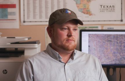 A man with a beard wearing a cap and light shirt sits indoors, with maps and a computer screen visible in the background.