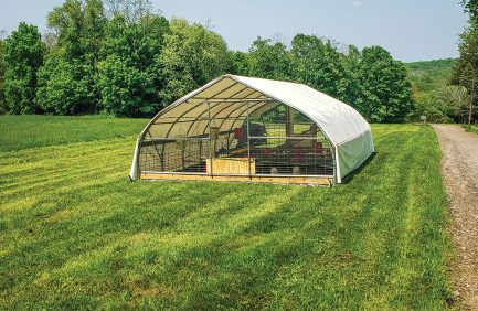 A large, white, hoop-style livestock shelter stands on a grassy field with trees in the background.