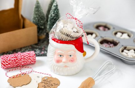 Santa Claus mug filled with wrapped treats, surrounded by baking items, red twine, gift tags, and small festive trees in the background.