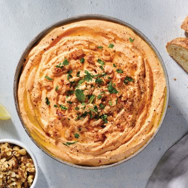 A bowl of creamy hummus garnished with herbs and spices, served with slices of bread and a small bowl of chopped nuts nearby.