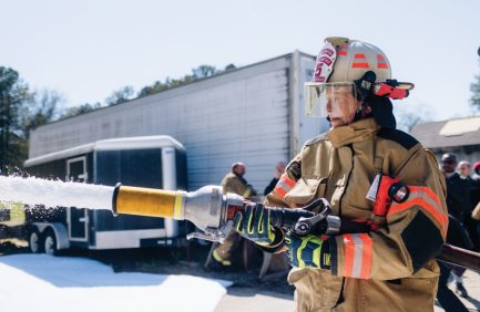 Firefighter in full gear operating a hose, spraying foam near trucks on a sunny day. Two other firefighters are visible in the background.