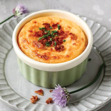 A baked savory soufflé in a green ramekin, topped with chopped herbs and crispy bits, placed on a decorative plate with purple flowers nearby.