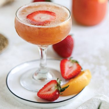 A cocktail in a coupe glass garnished with a strawberry slice, served on a plate with fresh strawberries and a lemon wedge.