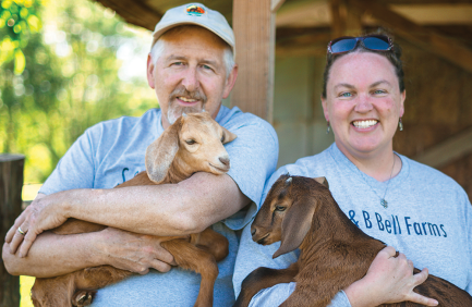 Two adults smiling outdoors, each holding a baby goat in their arms, standing close together in front of a wooden structure.