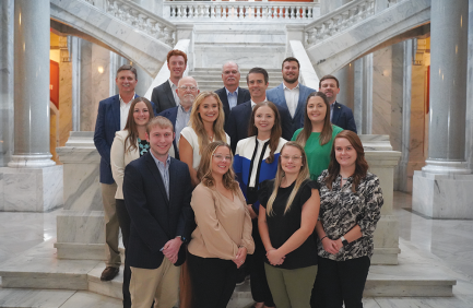 A group of people posing together on marble stairs inside a grand building with ornate railings and columns, dressed in business or business-casual attire.