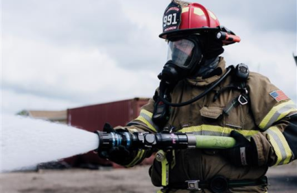 A firefighter in full gear sprays water from a hose, wearing a red helmet and protective mask, with shipping containers in the background.