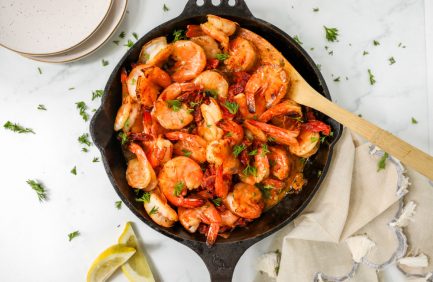 Shrimp sautéed with spices and herbs in a cast iron skillet, garnished with fresh parsley, with lemon wedges and plates nearby.