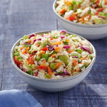 A bowl of colorful vegetable coleslaw with shredded cabbage, carrots, peppers, and onions on a wooden table, with another bowl in the background.