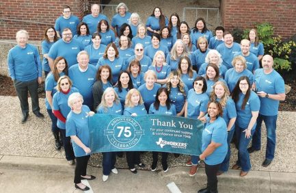 A large group of people in matching blue shirts stand outside a building, holding a banner celebrating a 75th anniversary and thanking supporters.