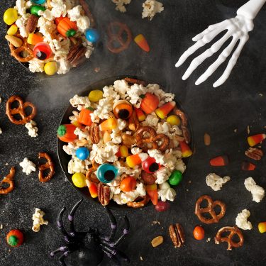 Halloween snack clusters with popcorn, pretzels, and colorful candy, surrounded by scattered snacks, a plastic spider, and a white skeleton hand on a dark surface.