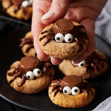 A hand holding a spider-themed cookie decorated with candy eyes and chocolate, with more similar cookies on a black plate.