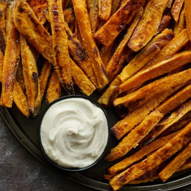 A plate of seasoned sweet potato fries served with a small bowl of creamy white dipping sauce.