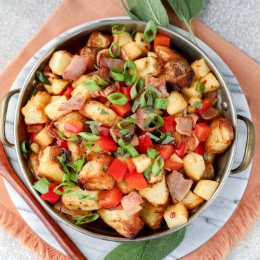 A bowl of roasted potatoes mixed with diced red bell peppers and garnished with chopped green onions, served on a round marble platter with fresh sage leaves.