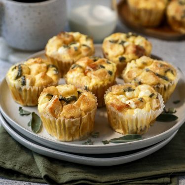 A plate of savory muffins with visible herbs and cheese, arranged on a white plate, set on a green napkin with a mug and more muffins in the background.