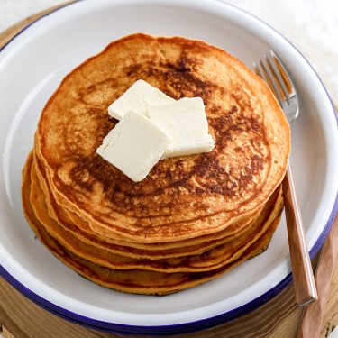A stack of golden brown pancakes topped with two pieces of butter, served on a white plate with a fork.