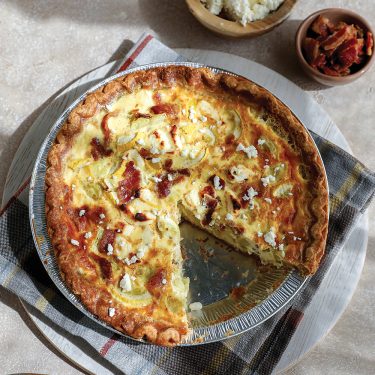 A baked quiche with a slice missing, placed on a cloth with small bowls of salad and a side dish nearby.