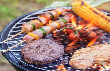 Grilled burger patty, chicken breast, skewers with vegetables and meat, whole tomatoes, and corn on the cob cooking over a charcoal barbecue.