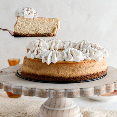 A creamy cheesecake topped with whipped cream, displayed on a cake stand, with one slice being lifted to show the texture.