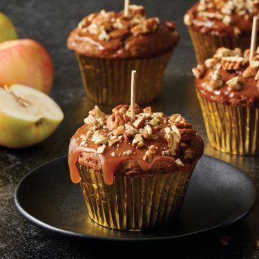 Caramel apple cupcakes with chopped nuts, topped with wooden sticks, sit on a black plate and dark surface, with apples in the background.