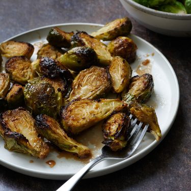 Roasted Brussels sprouts served on a white plate with a fork, with a side bowl of green vegetables in the background.