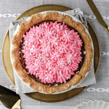 A pie with a golden crust, topped with pink whipped cream or frosting, on a decorative plate with cutlery beside it.