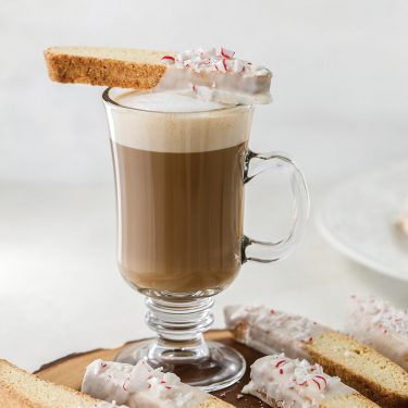 A glass mug of coffee with frothy milk, garnished with a biscotti topped with white icing and crushed peppermint, set on a wooden platter.