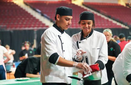 Two chefs in white uniforms and black hats prepare food together in a large indoor setting, smiling and focused on their task.