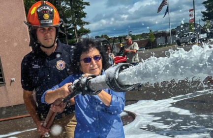 A firefighter assists a woman spraying water from a fire hose outdoors, with foam and people visible in the background.
