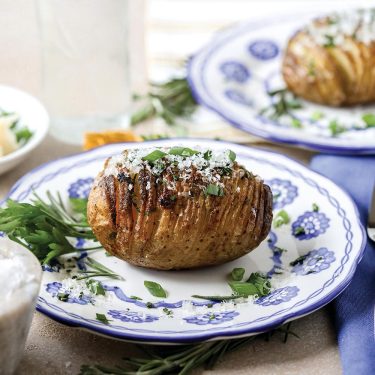 A baked Hasselback potato garnished with herbs and cheese, served on a decorative blue and white plate with fresh greens.