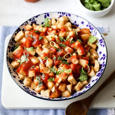 A bowl of diced potatoes topped with tomato sauce and chopped herbs, served on a white plate with a wooden spoon beside it.