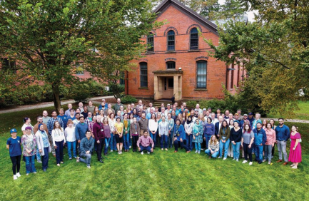 A large group of people poses for a photo on green grass in front of a brick building with arched windows and trees.
