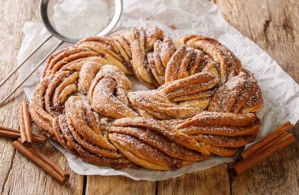 A braided cinnamon bread wreath dusted with powdered sugar sits on parchment paper atop a rustic wooden surface.