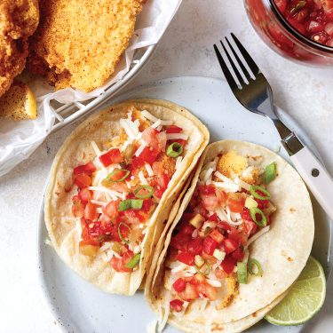Two soft tacos filled with breaded fish, diced tomatoes, shredded cheese, and green onions, served with lime wedges on a white plate.
