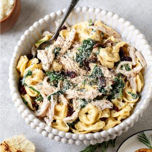 Bowl of tortellini pasta topped with shredded chicken, creamy sauce, herbs, and pepper, with a fork inside the bowl.