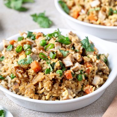 A bowl of fried rice with diced vegetables, peas, carrots, tofu, and herbs, served in a white bowl on a table.
