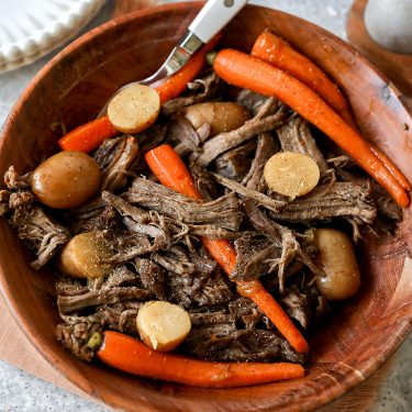 A bowl of pot roast with shredded beef, carrots, potatoes, and round slices of parsnip, served on a wooden plate with a fork.