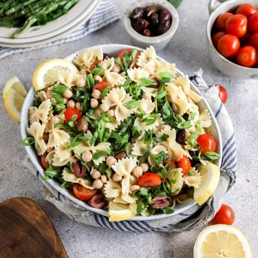 Bowl of pasta salad with cherry tomatoes, white beans, olives, fresh herbs, and lemon slices, surrounded by small bowls of olives, tomatoes, and greens.