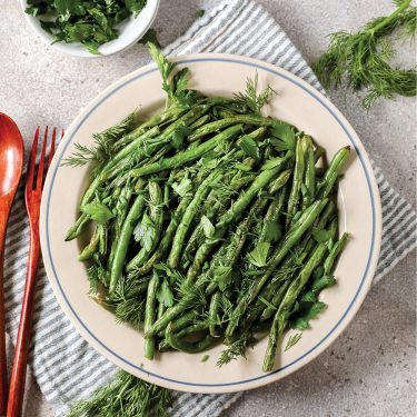 A bowl filled with cooked green beans garnished with fresh herbs, placed on a striped cloth with utensils and a bowl of extra herbs nearby.