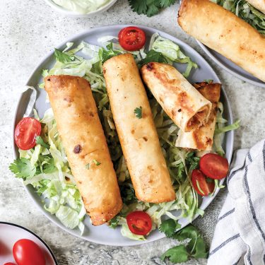 Three crispy taquitos served on a bed of lettuce and sliced cherry tomatoes, presented on a round plate with fresh herbs.